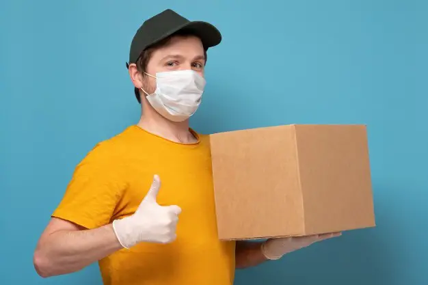 smiling young delivery man holding card box wearing mask