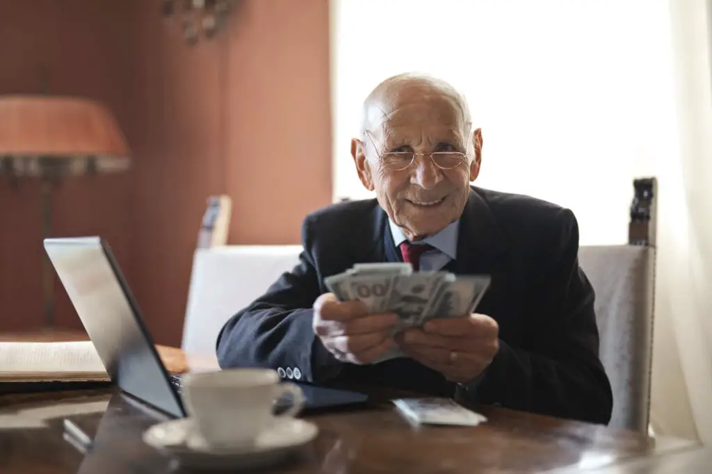 confident-senior-businessman-holding-money-in-hands-while-sitting-at-table-near-laptop